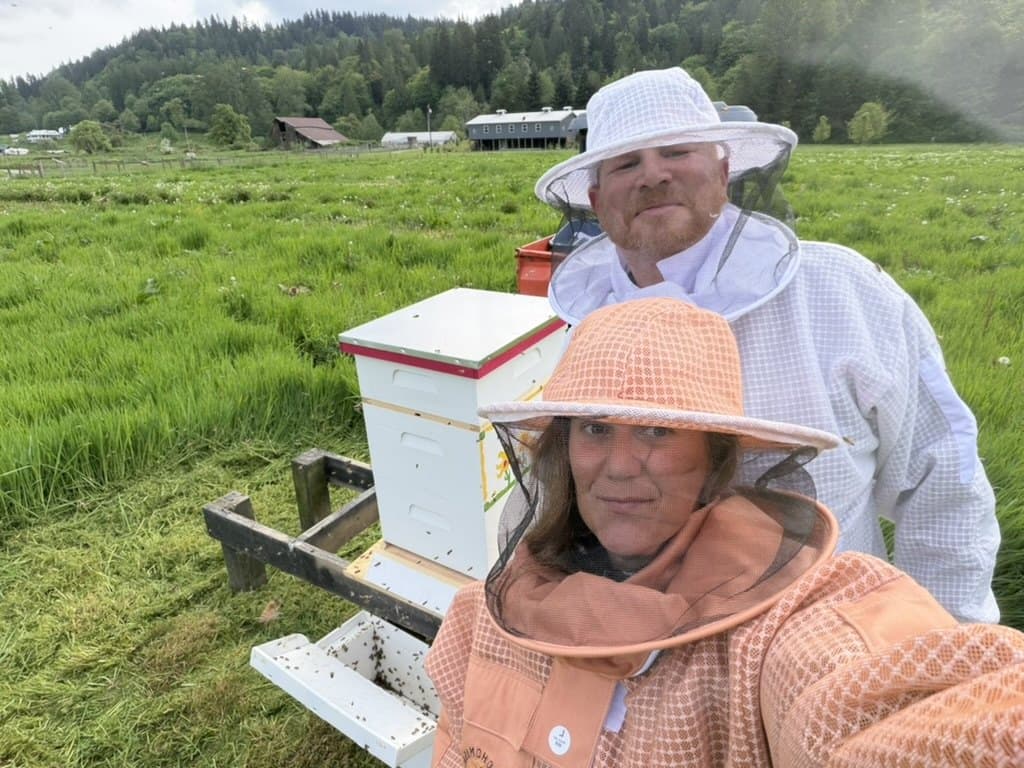 Kappie Ayers and Joe Ayers in beekeeping suits next to one of the hives at Wild Roots, with the pasture and a barn in the distance